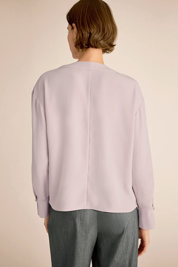 Person wearing a light purple blouse and gray pants on a beige background | Exclude | Grid Hover