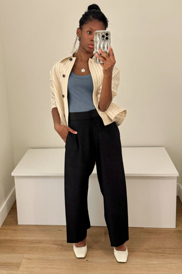 Person taking a mirror selfie wearing a beige cardigan, blue top, black pants, and white shoes in a minimalistic room. | Exclude