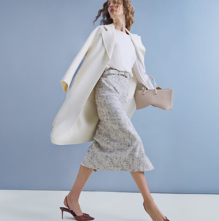 Woman in a white coat and patterned skirt holding a straw bag against a light blue background