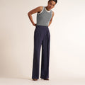 A model wearing high-waisted navy blue wide-leg trousers with pleats and a gray sleeveless knit top with a cream collar, standing against a solid off-white studio background. | Lead