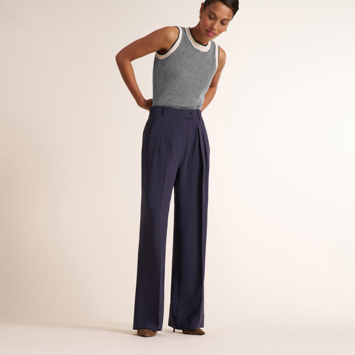 A model wearing high-waisted navy blue wide-leg trousers with pleats and a gray sleeveless knit top with a cream collar, standing against a solid off-white studio background. | Lead