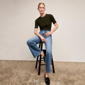 Person wearing a black t-shirt and blue jeans sitting on a stool against a plain background | Lead