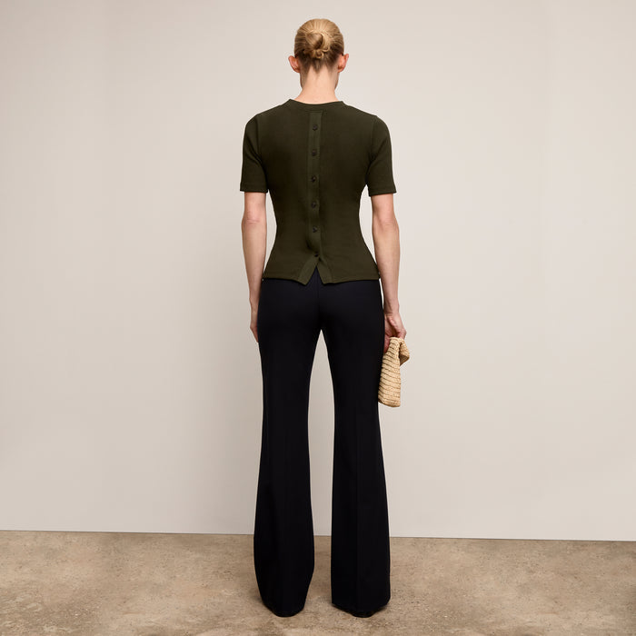 Person wearing a green top and black pants holding a straw bag against a plain background