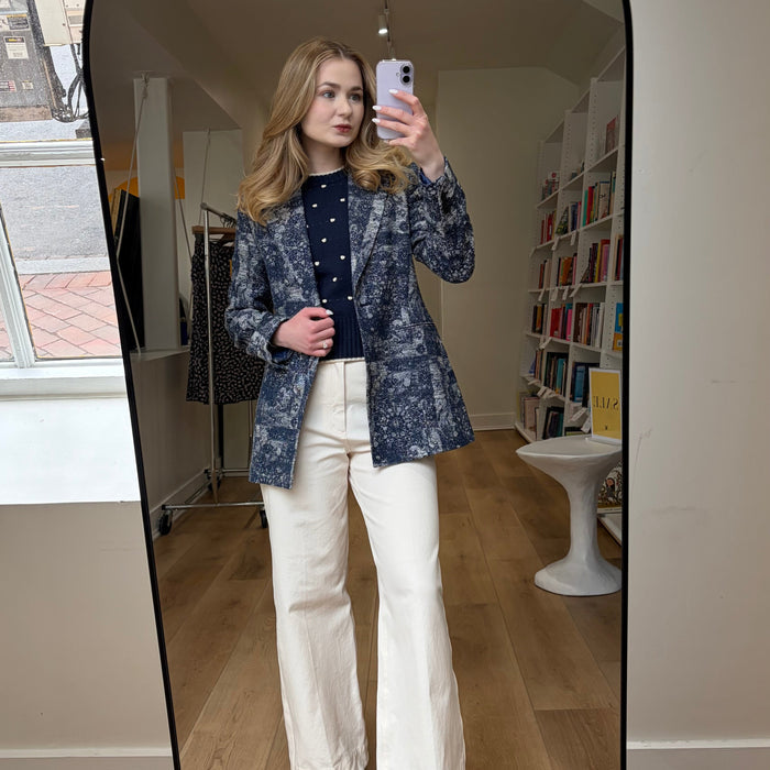 Person taking a mirror selfie wearing a patterned blazer and white pants in a room with books and furniture.