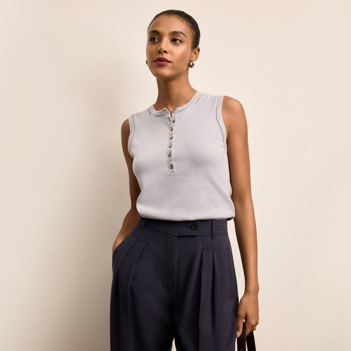 Woman wearing a light gray sleeveless top with buttons and dark blue pants against a beige background | Lead