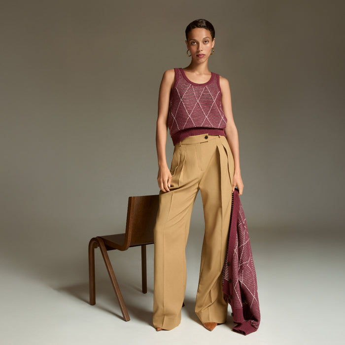 Woman wearing a patterned sleeveless top and beige pants, holding a matching fabric, against a neutral background.