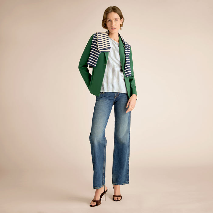Woman wearing a green blazer, striped shirt, and blue jeans on a beige background