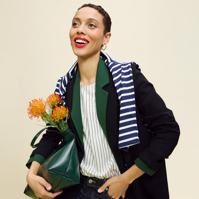 Woman holding a green handbag and flowers, wearing a black coat with a striped scarf on a beige background