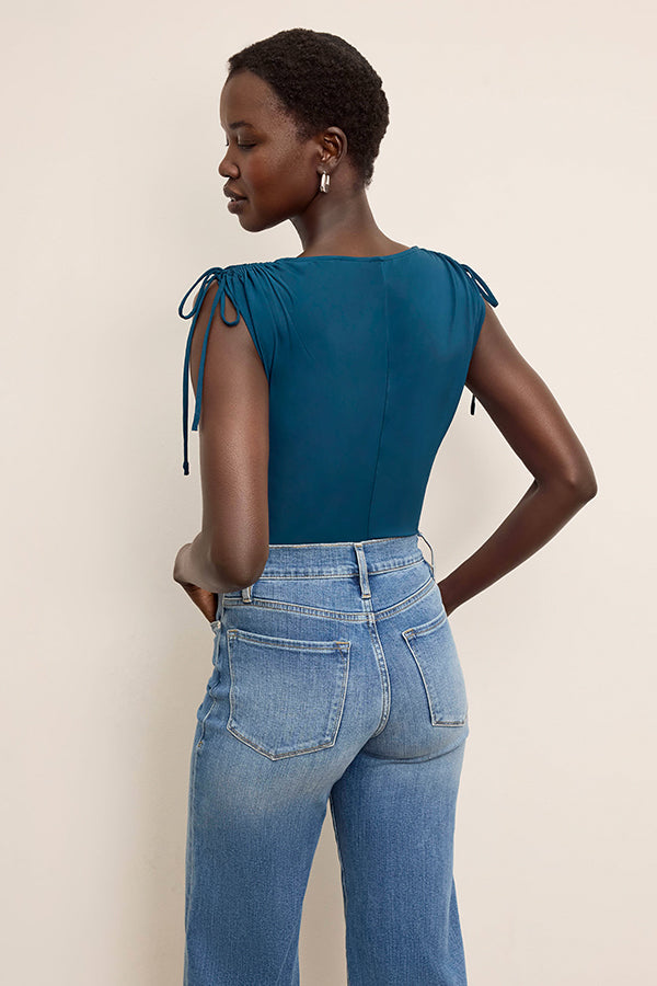 Woman wearing a teal top with tie sleeves and blue jeans against a beige background | Exclude