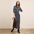 Woman wearing a navy dress with white floral pattern, holding a brown handbag against a beige background | Lead