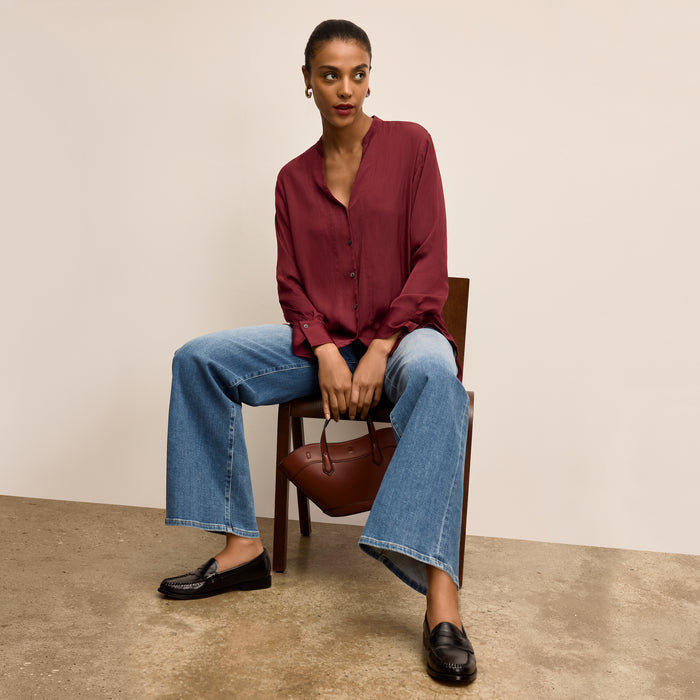 Woman sitting on a chair holding a brown handbag, wearing a red blouse and blue jeans | Lead