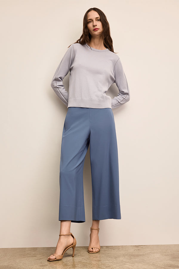 Woman wearing a light gray top and blue pants against a beige wall. | Exclude | Grid
