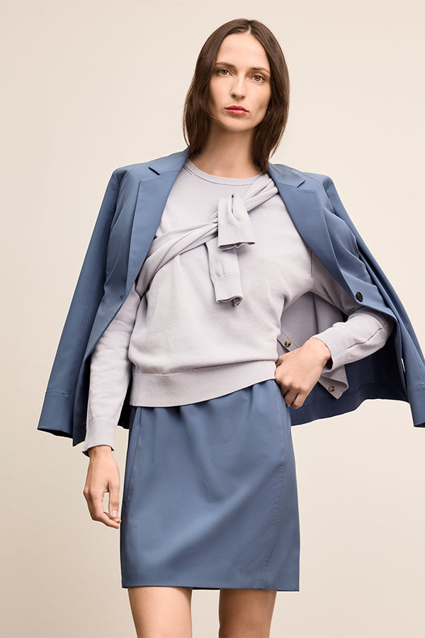 Woman wearing a light gray sweater with a bow, blue skirt, and blue blazer on a beige background | Exclude