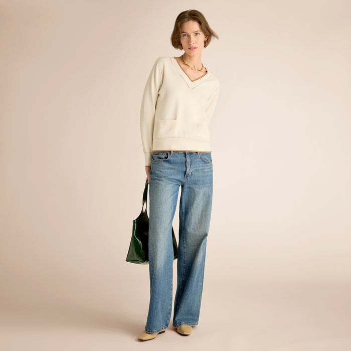 Woman wearing a white sweater and blue jeans holding a green bag on a beige background | Lead