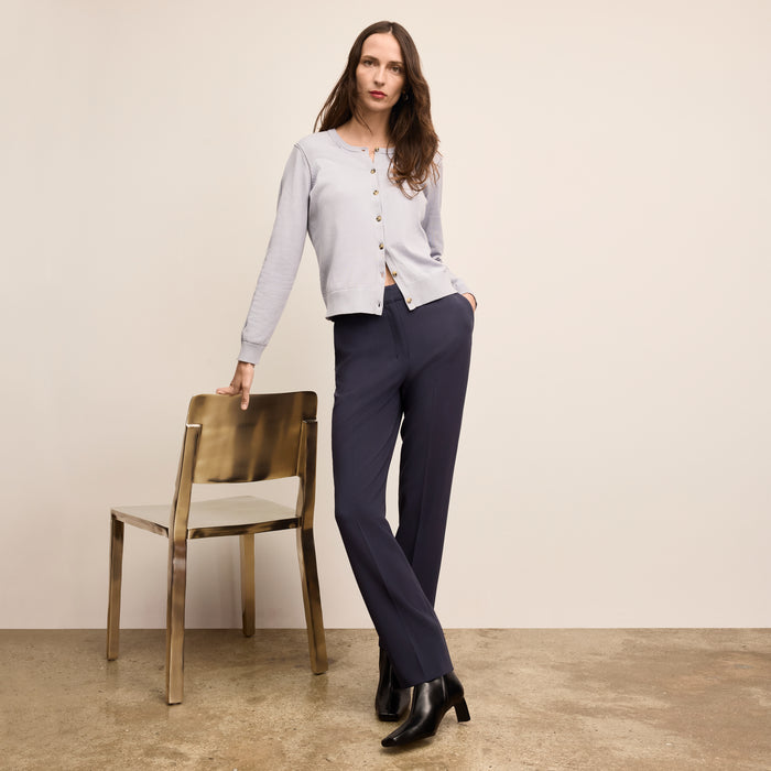Woman in a light gray cardigan and navy pants standing next to a gold chair against a beige wall. | Lead