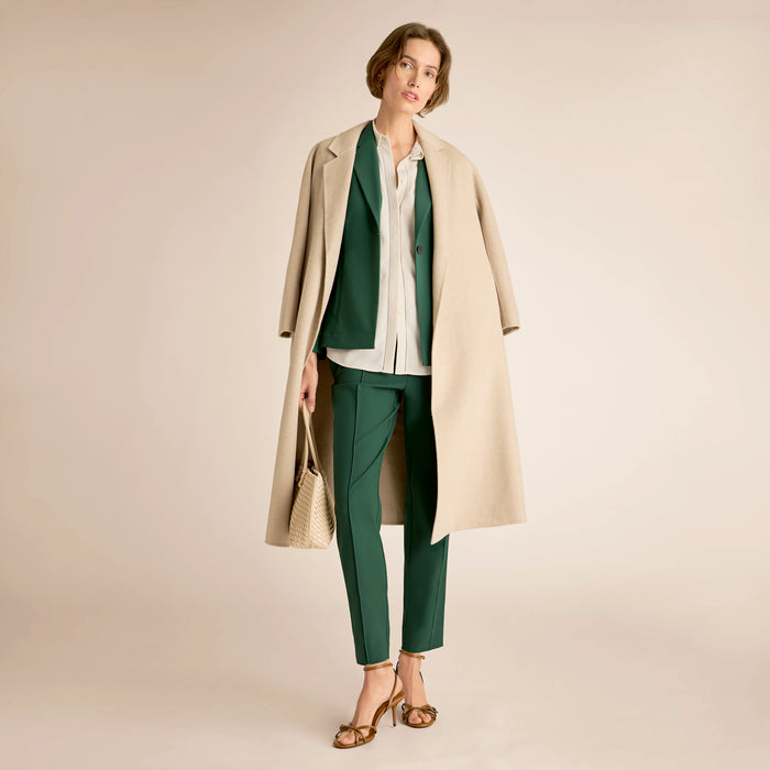 Woman wearing a beige coat, green pants, and white shirt on a beige background