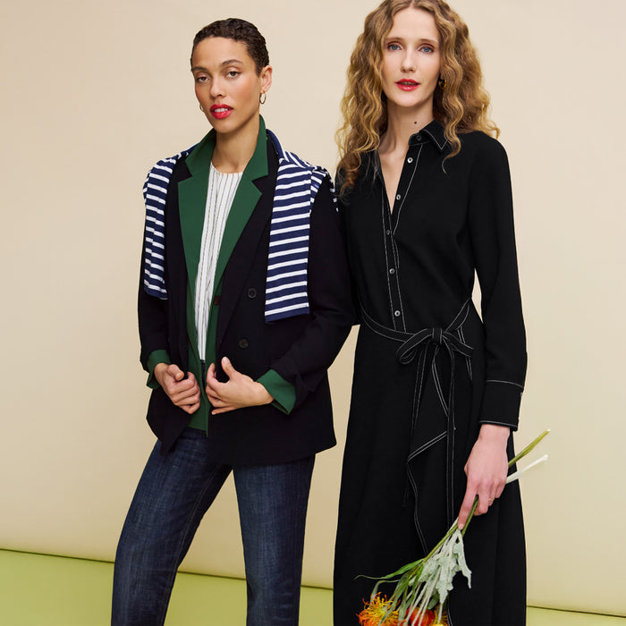 Two women standing side by side against a beige background, one wearing a striped blazer and the other in a black dress with a belt.