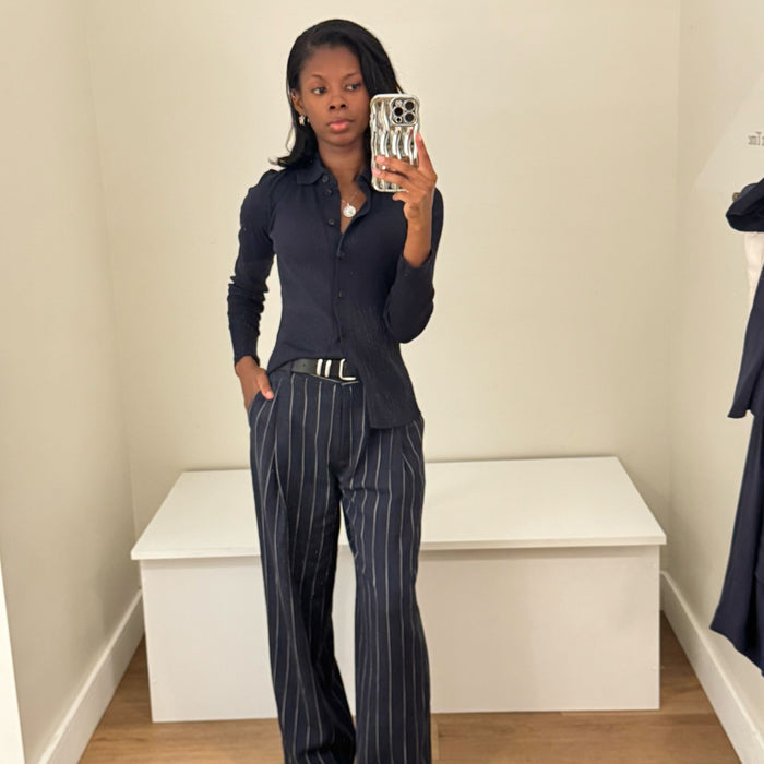 Person taking a mirror selfie wearing a dark blue shirt and pinstripe pants in a minimal room.