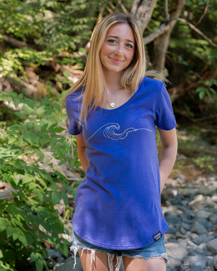 A woman with blonde hair stands outdoors in a wooded, rocky area wearing a blue short-sleeve t-shirt with a white wave graphic on the front and denim shorts.