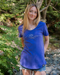 A woman with blonde hair stands outdoors in a wooded, rocky area wearing a blue short-sleeve t-shirt with a white wave graphic on the front and denim shorts.