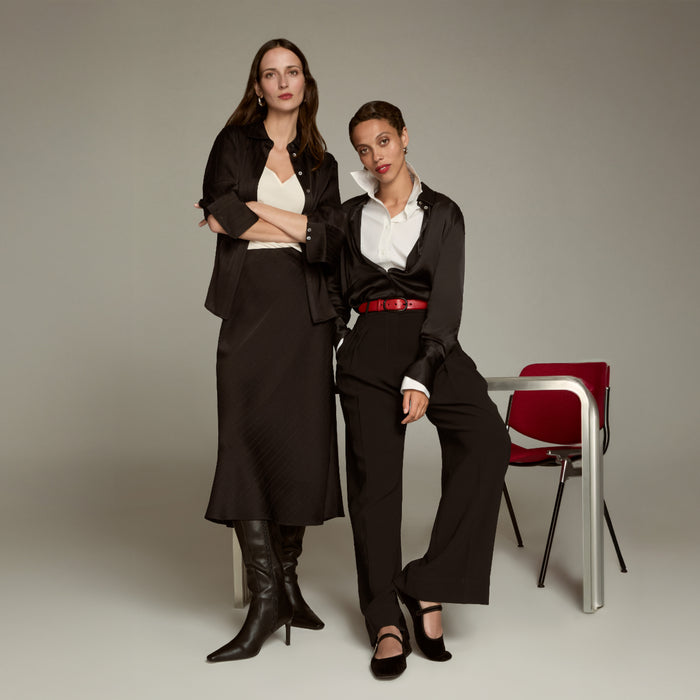 Two women in professional attire standing in a studio setting with a chair in the background.