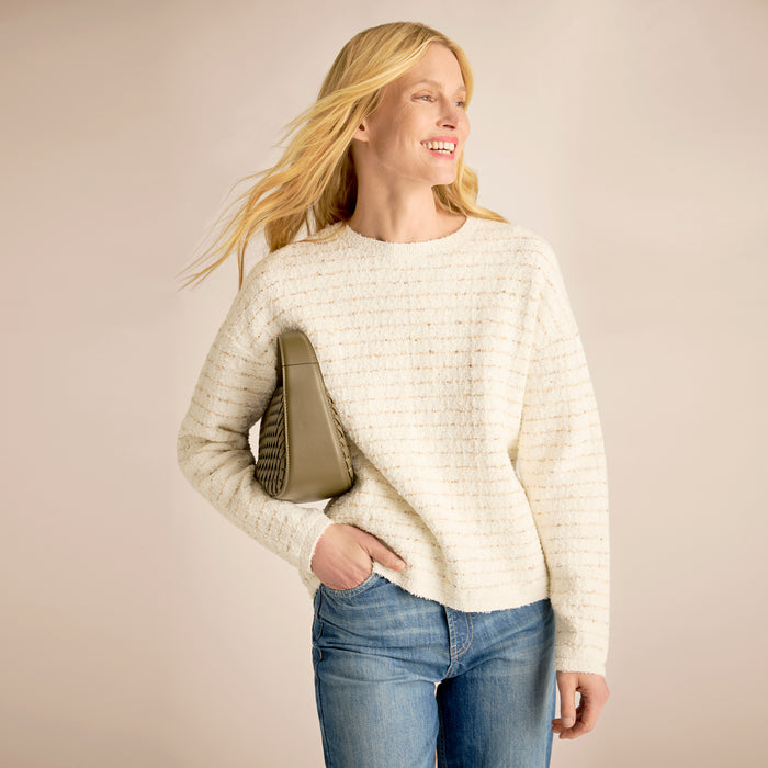 Woman wearing a cream sweater and blue jeans, holding a beige handbag against a plain background | Lead