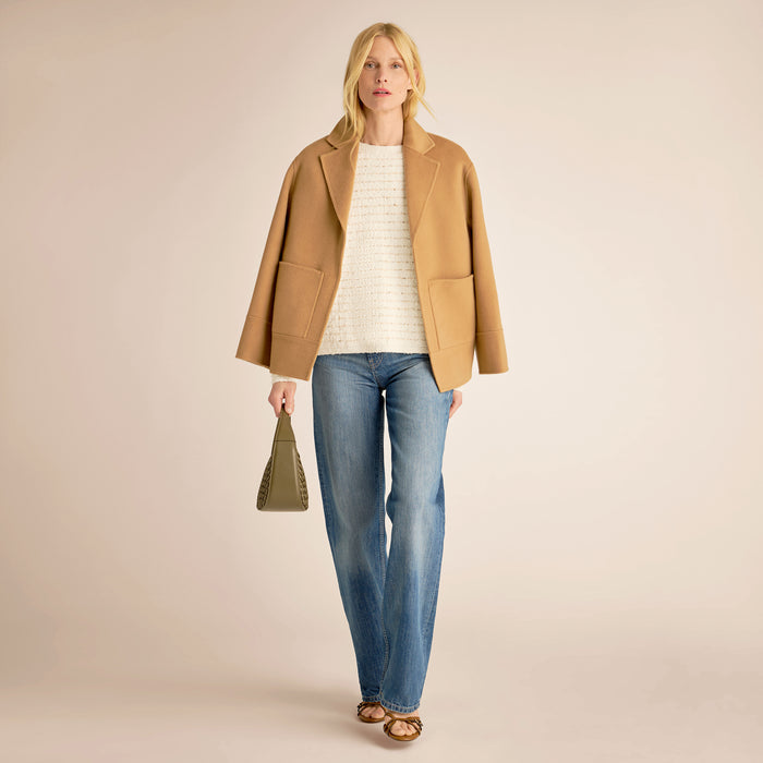 Woman wearing a tan coat, white sweater, blue jeans, and holding a green bag on a beige background