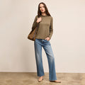 Woman wearing a green sweater and blue jeans holding a woven bag against a beige background | Lead