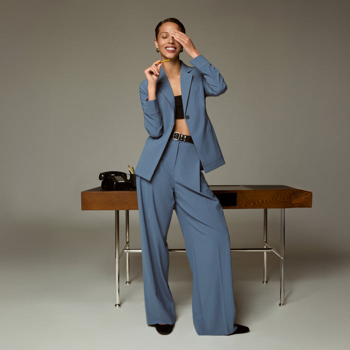 Woman in a blue suit standing next to a wooden desk with a phone on it.