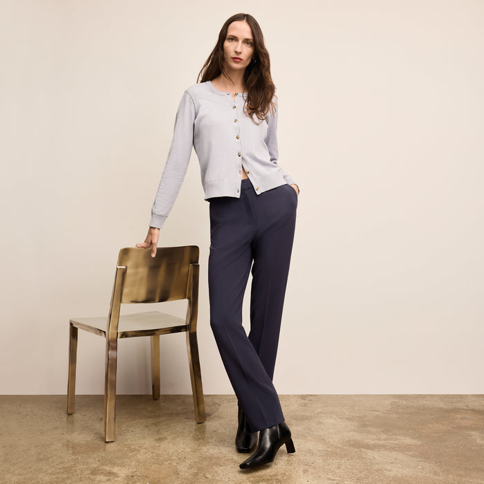 Woman in a light gray cardigan and navy pants standing next to a gold chair against a beige wall.