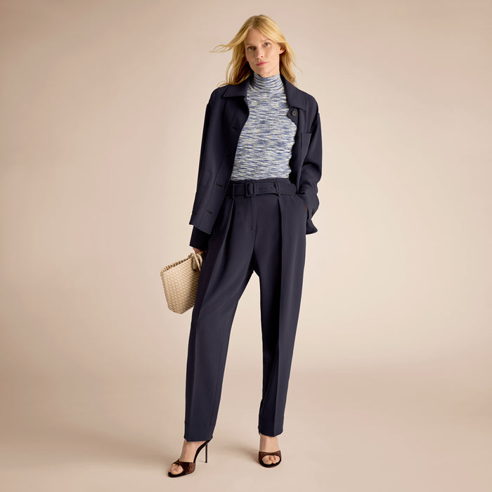 Woman wearing a navy suit with a light gray turtleneck and holding a straw bag on a beige background