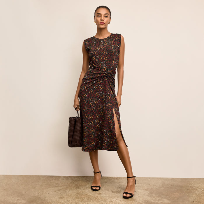 Woman wearing a patterned dress with a slit, holding a matching handbag against a beige background. | Lead
